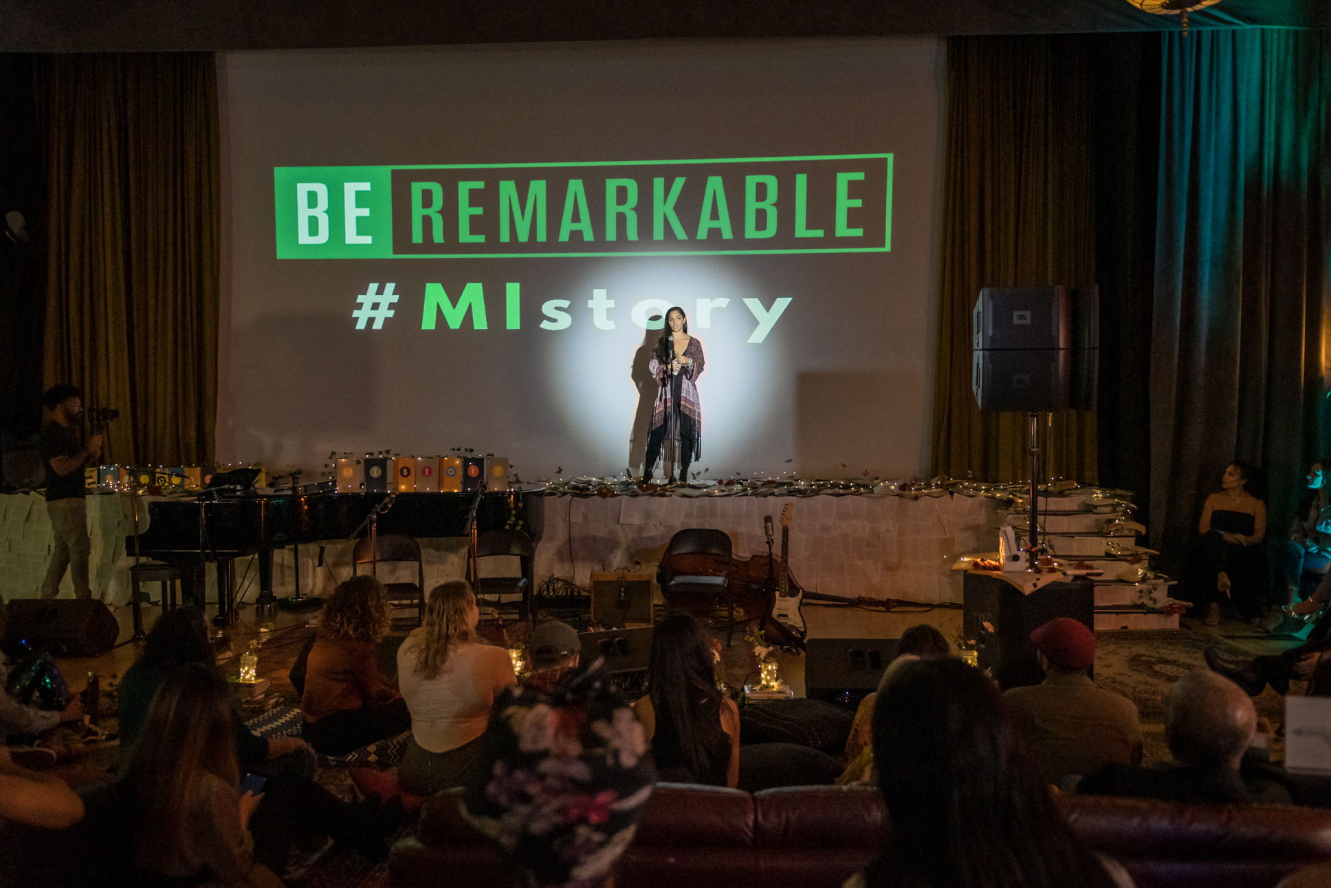 MIstory in Los Angeles &mdash; the BE REMARKABLE / #MIstory projection glowing over the stage as a teen storyteller takes the floor among lit candles, a piano, and an intimate audience seated on rugs.